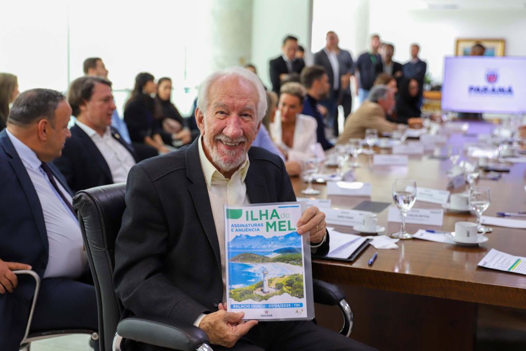 Novo marco regulatório da Ilha do Mel avança com instituição de comitê gestor 3 Imagem de um evento em que um homem sorridente segura uma revista sobre a Ilha do Mel, com empresários e autoridades ao fundo em uma mesa de reuniões. Novo marco regulatório da Ilha do Mel avança com instituição de comitê gestor. Novo marco regulatório da Ilha do Mel avança com instituição de comitê gestor