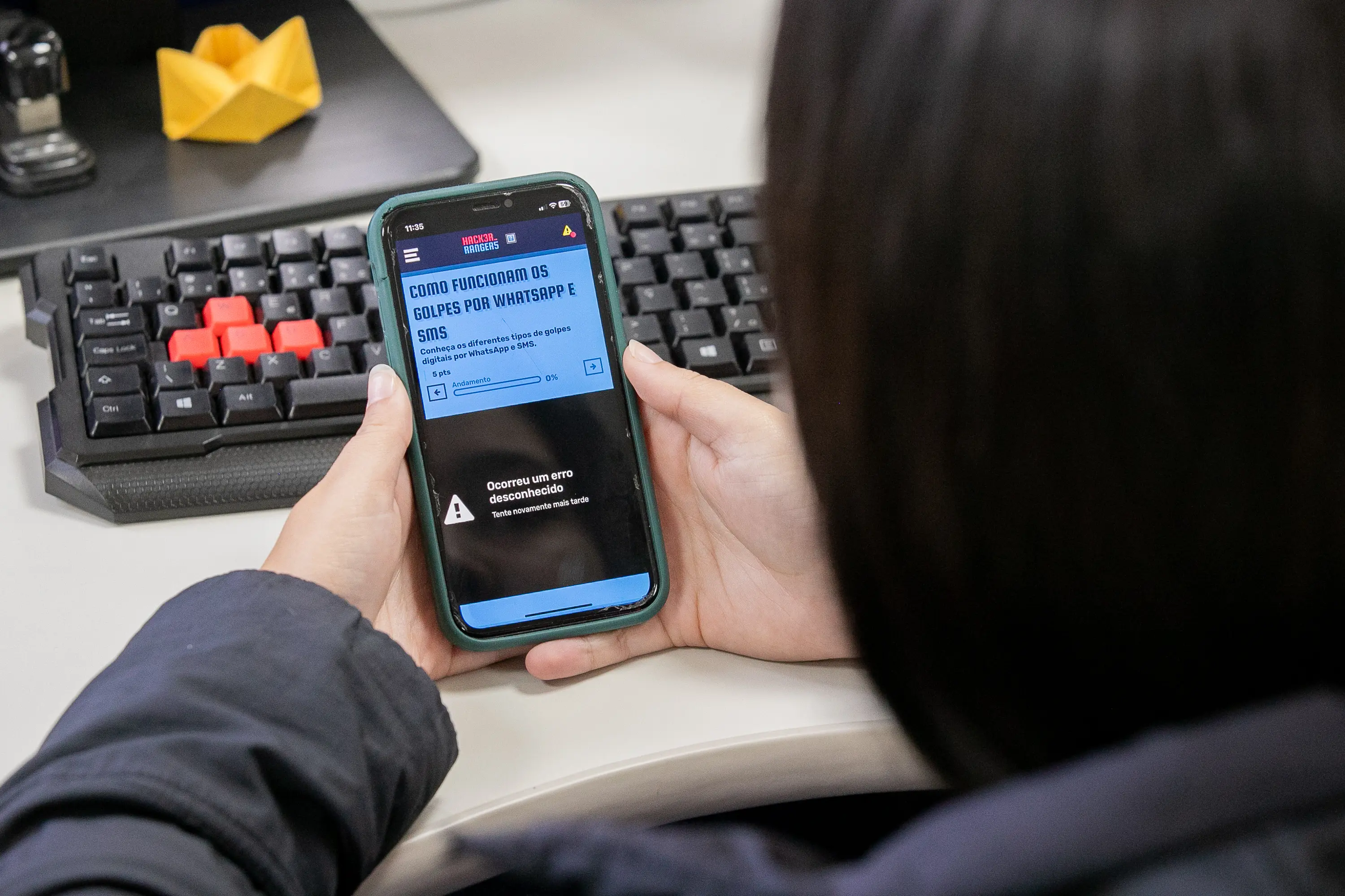 Pessoa usando smartphone para ler mensagem de aviso sobre golpes por WhatsApp e SMS, com teclado de computador ao fundo e detalhes de escritório. Hacker Rangers entra na quinta temporada com novidades