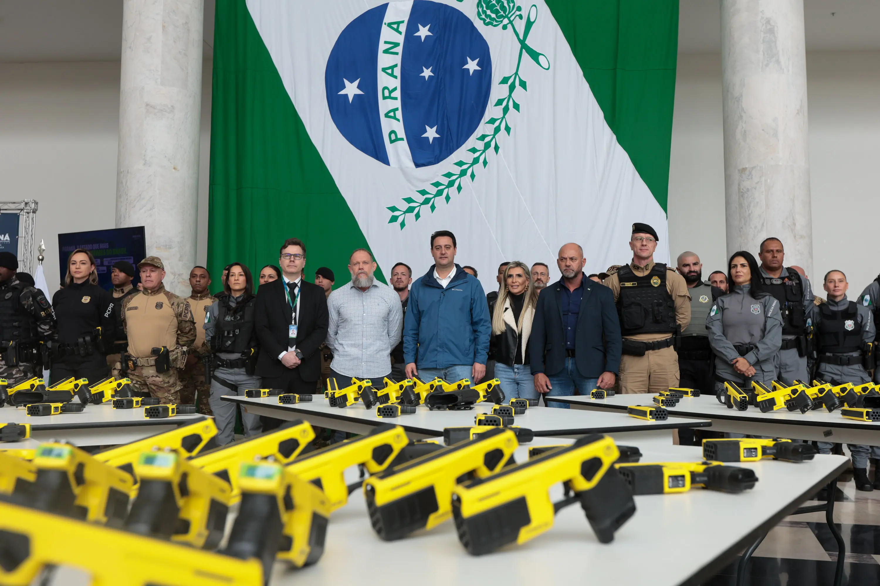 Autoridades e policiais de Parana participaram de uma cerimônia de entrega de armas, incluindo pistolas amarelas, com a bandeira do Paraná ao fundo, em um evento oficial. Com tecnologia de última geração, Paraná entrega 1,4 mil armas não letais para as polícias