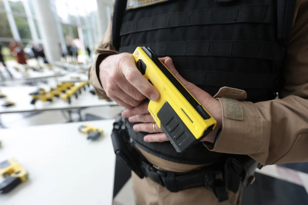 Com tecnologia de última geração, Paraná entrega 1,4 mil armas não letais para as polícias 3 Policial segurando uma pistola de tinta amarela em um ambiente com várias ferramentas à mesa ao fundo, representando ferramentas para aplicação de tinta ou marcações de segurança. Com tecnologia de última geração, Paraná entrega 1,4 mil armas não letais para as polícias