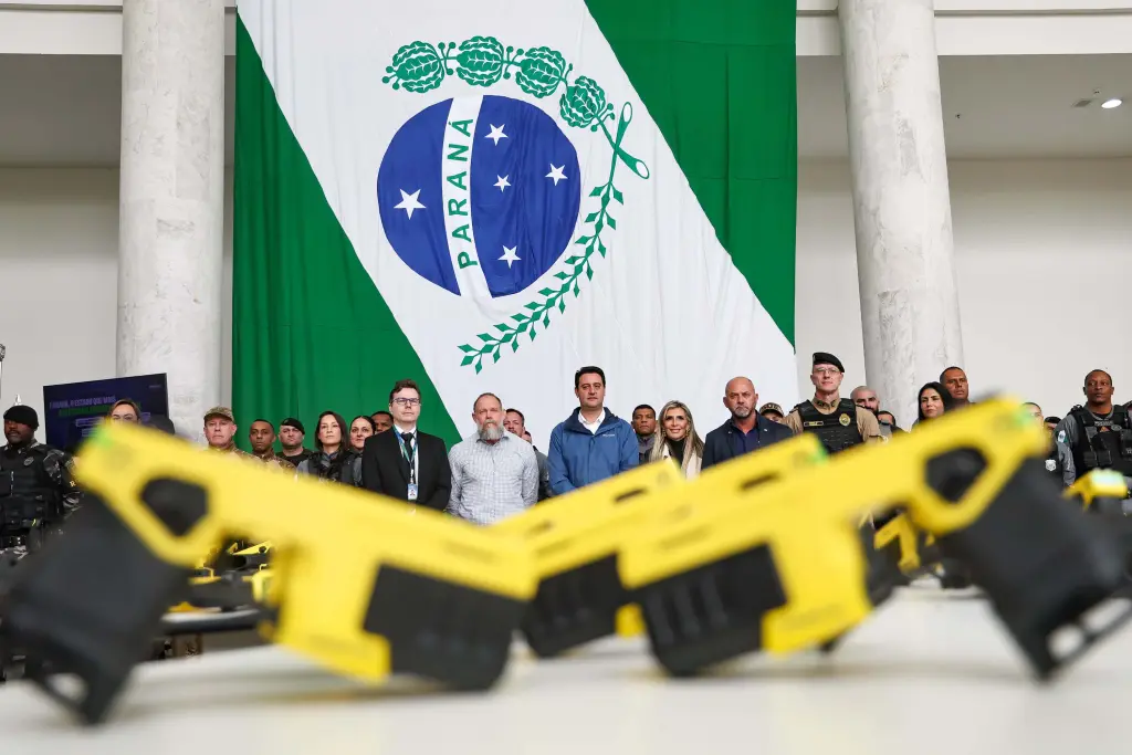 Com tecnologia de última geração, Paraná entrega 1,4 mil armas não letais para as polícias 2 Reunião oficial com representantes governamentais e policiais diante da bandeira de Paraná, destaque para armas de brinquedo na frente e grupo diverso ao fundo. Com tecnologia de última geração, Paraná entrega 1,4 mil armas não letais para as polícias