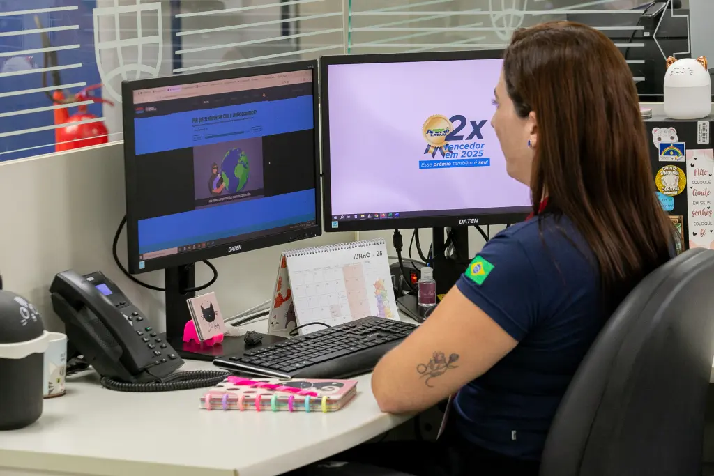 Hacker Rangers entra na quinta temporada com novidades 2 Mulher com tatuagem no braço, vestindo camisa azul com a bandeira do Brasil, trabalhando em um escritório com dois monitores, um calendário e objetos de decoração ao redor, em um ambiente de trabalho. Hacker Rangers entra na quinta temporada com novidades