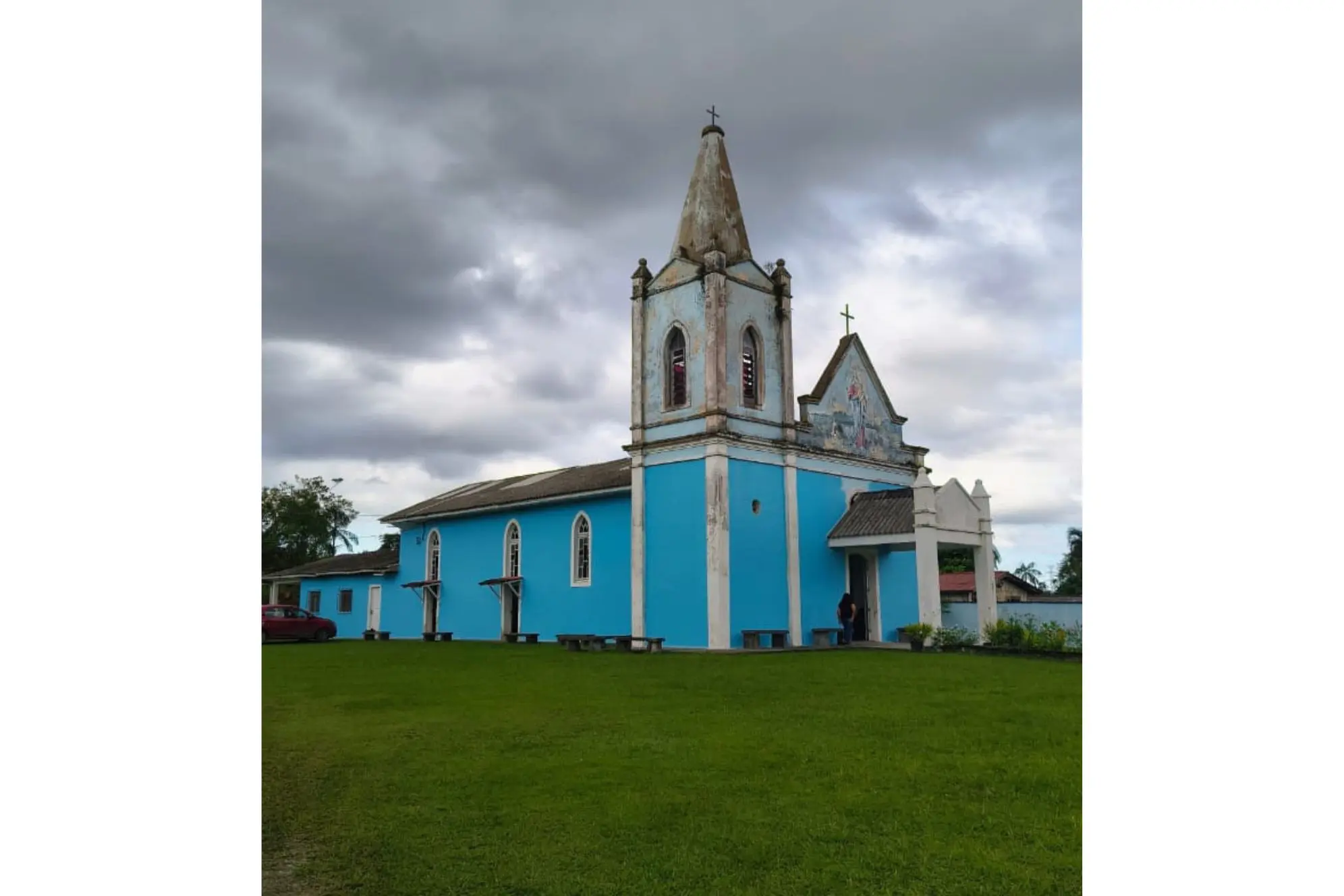 Igreja azul com detalhes arquitetônicos em um fundo de céu nublado. A edificação destaca-se na paisagem rodeada por gramado verde. Bazar Solidário do Instituto Palazzolo será realizado neste sábado na Comunidade Nossa Senhora Auxiliadora