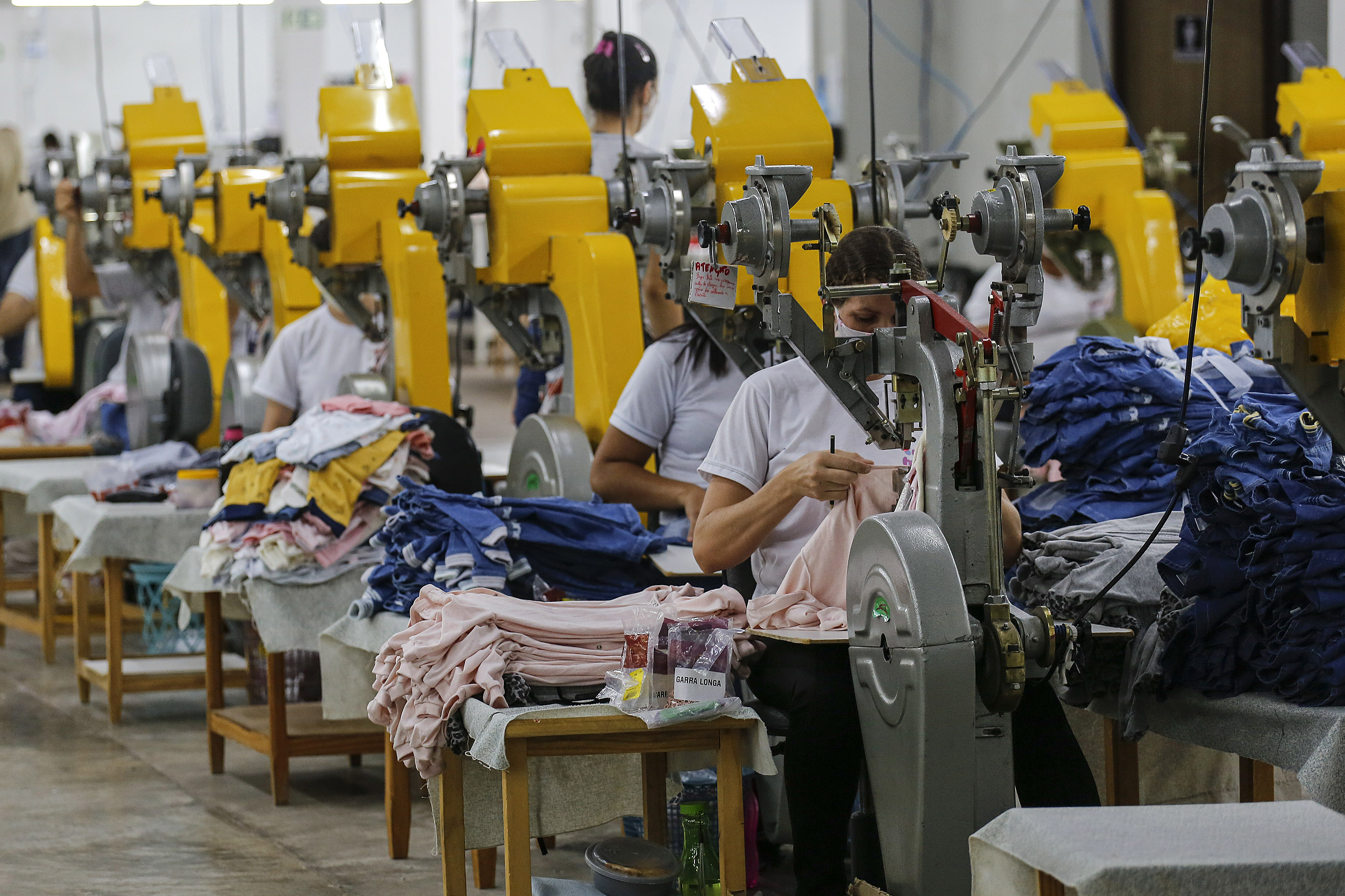 Imagem de uma fábrica de confecção com trabalhadores em máquinas de costura, mostrando a produção de roupas com tecidos em mesas ao redor. Ambiente iluminado e organizado. 5.º melhor do País: Paraná gerou 60,7 mil empregos de janeiro a março de 2025