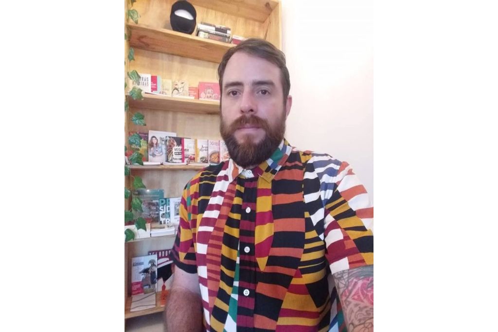 Mutirão Artístico: Paranaguá Mais Cores revitalizará pista de skate 1 Homem com uma camisa estampada colorida em um ambiente interno, com prateleiras ao fundo cheias de livros e itens decorativos. Mutirão Artístico: Paranaguá Mais Cores revitalizará pista de skate