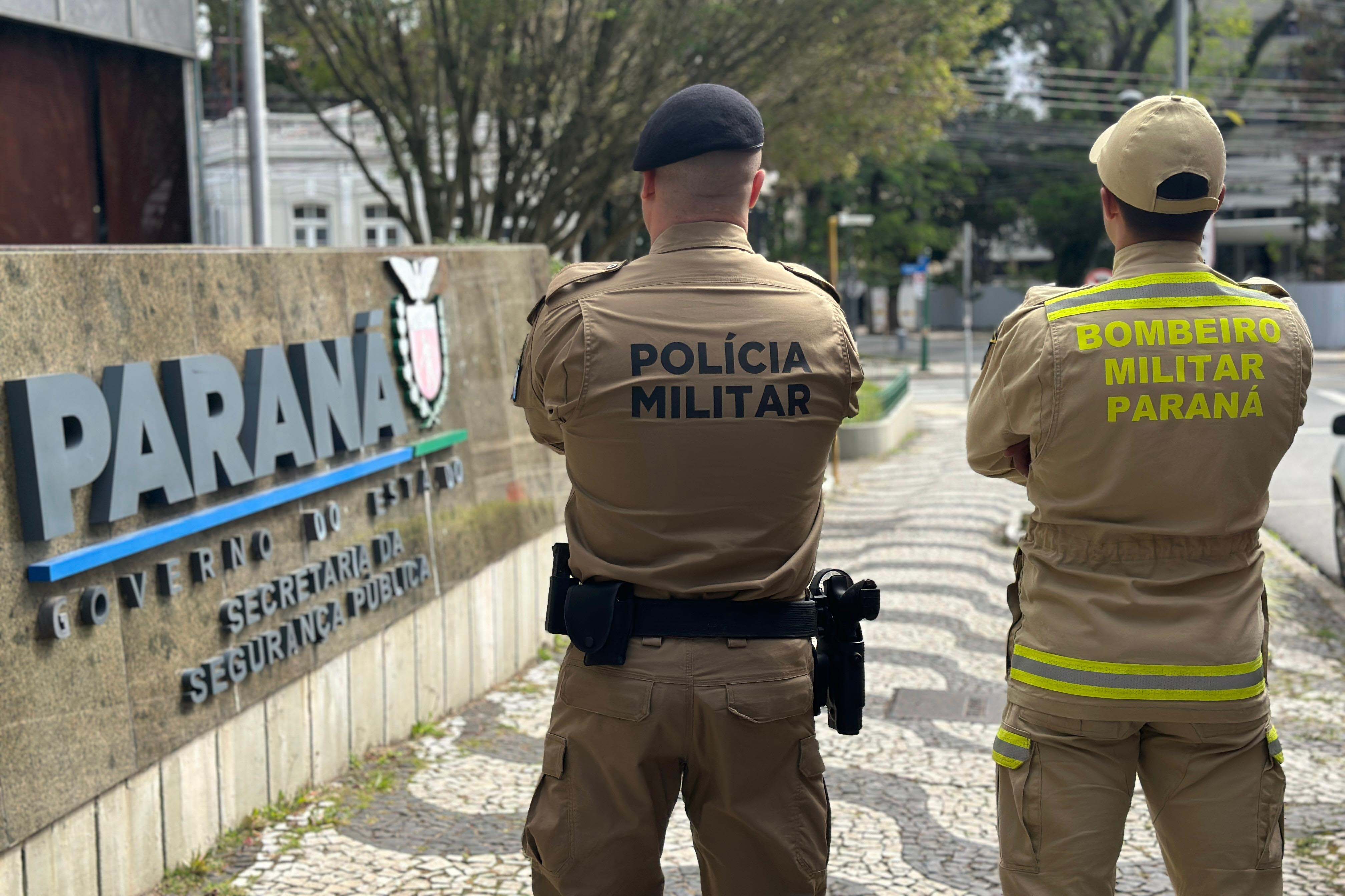 Policiais militares do Paraná em frente à sede do governo estadual, com destaque para a sinalização do Paraná na parede de concreto, em uma cena urbana com árvores e edifícios ao fundo. Concurso público da PMPR e Corpo de Bombeiros tem mais de 30,3 mil inscritos
