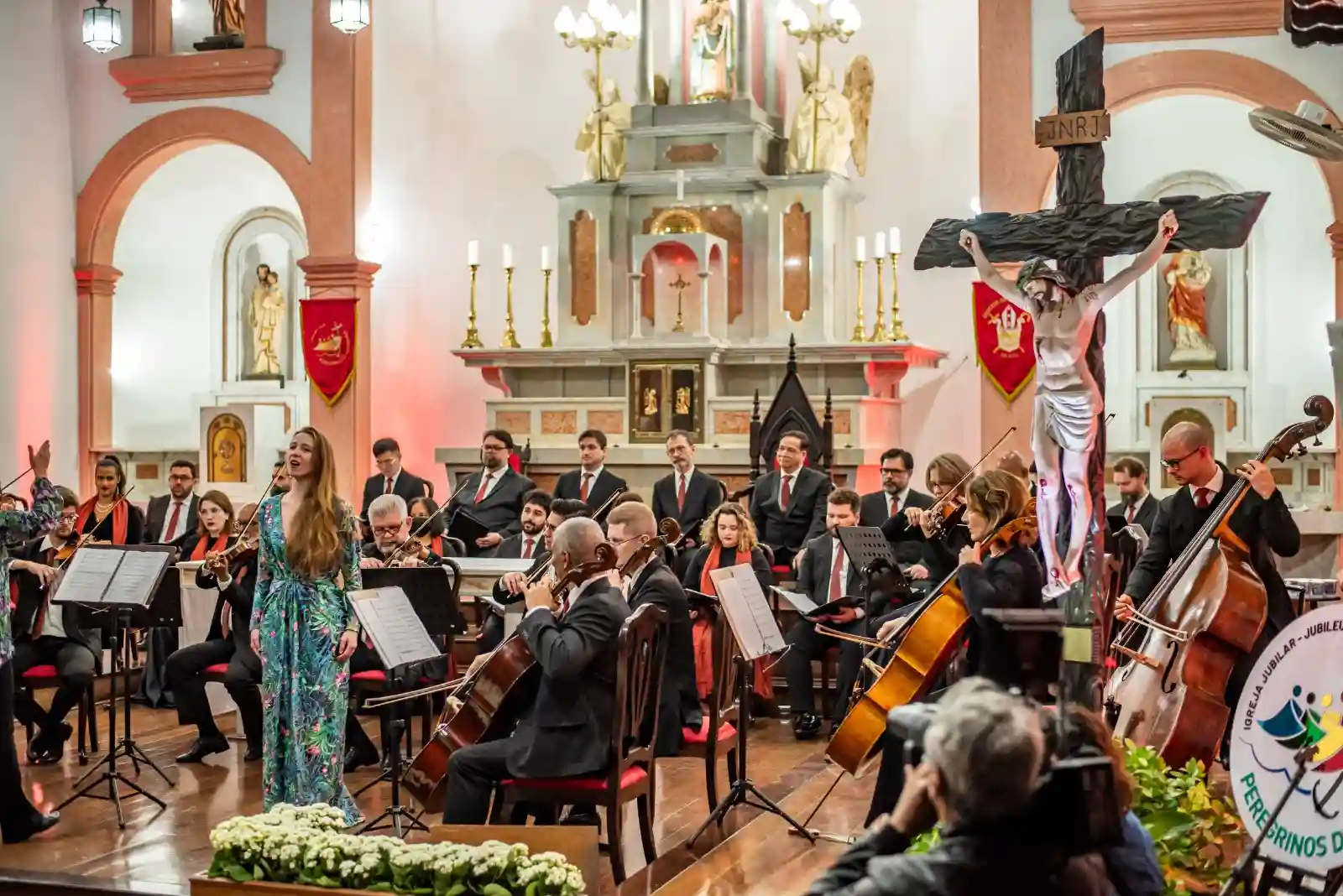 Camerata Antiqua de Curitiba se apresenta em Paranaguá, com músicos tocando instrumentos clássicos e uma cantora em um palco de igreja, acompanhada por uma cruz e elementos religiosos.