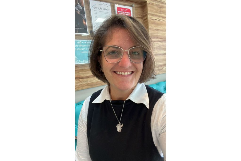 Mulher sorridente usando óculos, camiseta branca, suéter preto e colar com pingente, sentada em ambiente interno com parede de madeira ao fundo. Nova Acrópole realiza palestra gratuita sobre “Como Superar Seus Limites Internos”