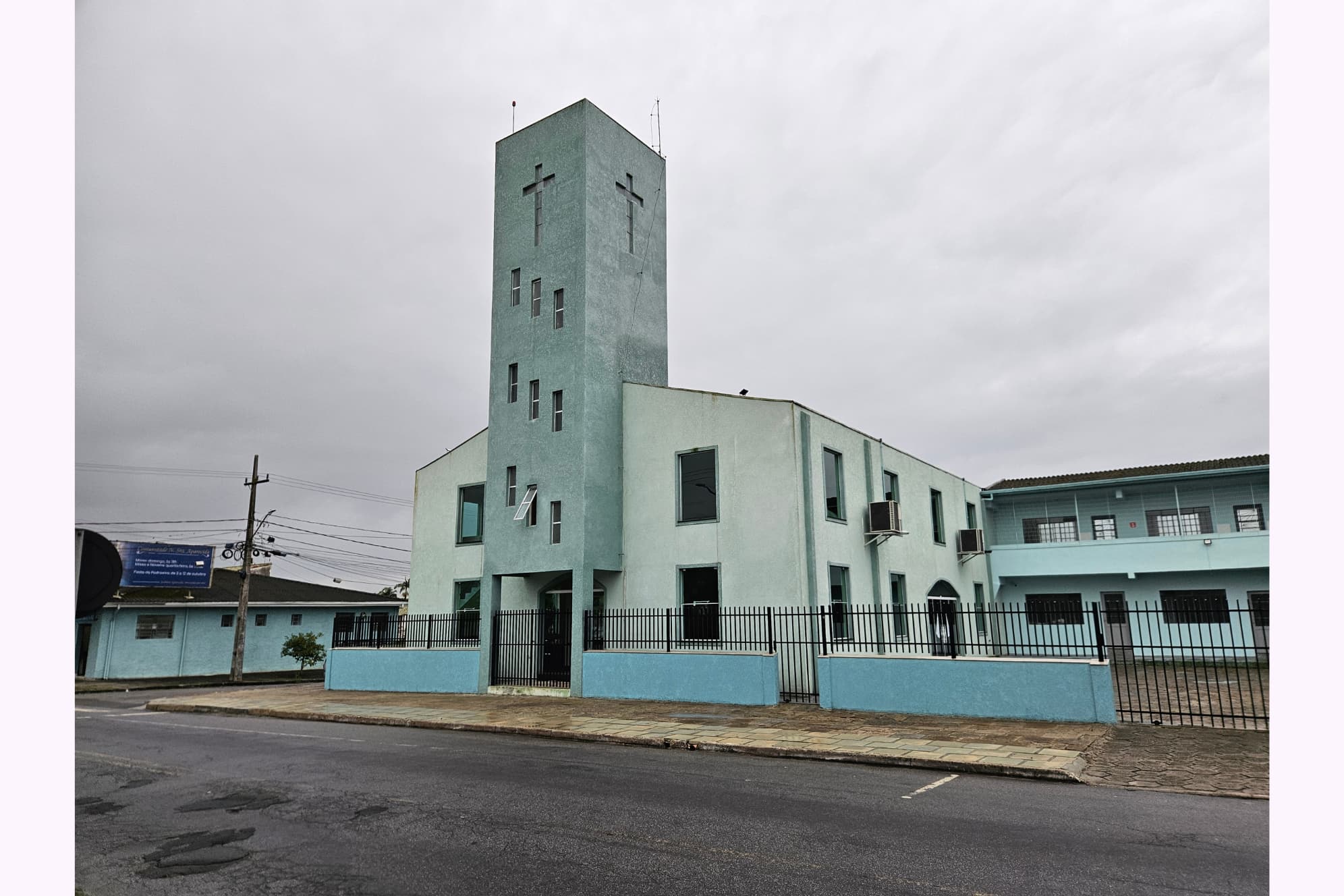 Edifício de igreja com torre e cruzes na fachada, localizado na cidade, sob céu nublado, com cerca de metal em primeiro plano, representando arquitetura religiosa. Festa e Novenário em honra a Nossa Senhora Aparecida começa nesta sexta-feira em Paranaguá