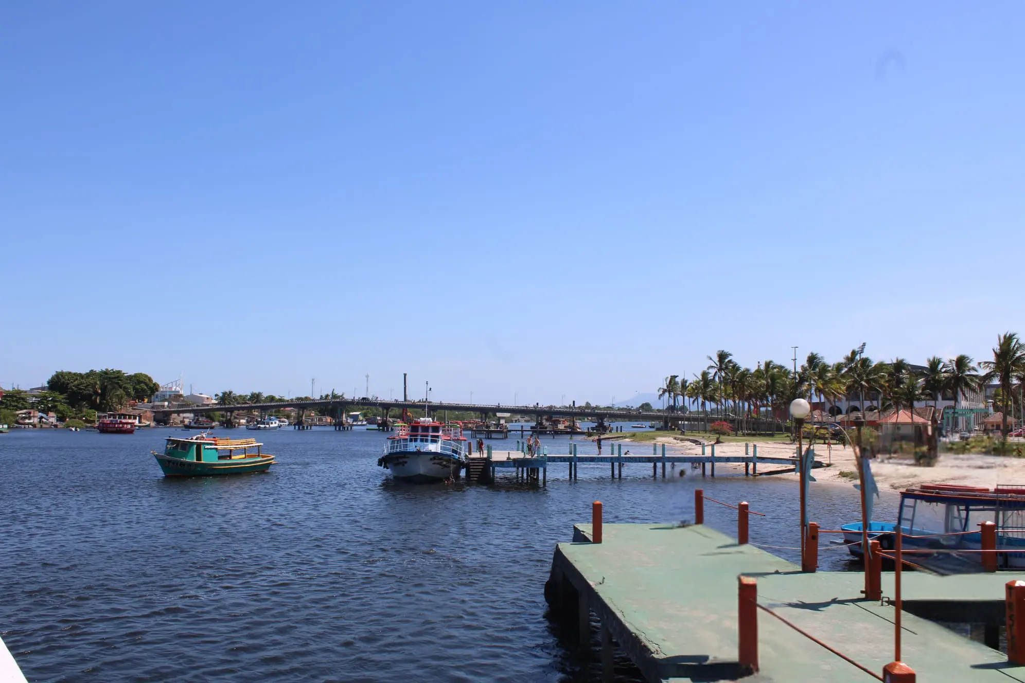 Cais de porto com barcos navegando em águas calmas e céu azul, ao fundo uma linha de coqueiros e uma ponte. Paisagem litorânea tranquila e ensolarada. Feriado de Nossa Senhora do Rosário: veja o que abre e o que fecha em Paranaguá nos dias 6 e 7 de outubro
