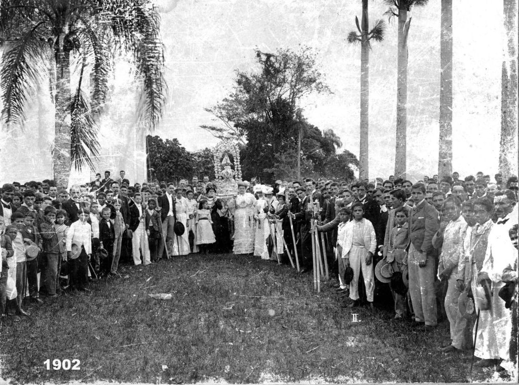 História da Festa de Nossa Senhora do Rocio: origem, tradição e fé do povo paranaense 2 Grupo grande de pessoas reunidas ao ar livre na procissão de Nossa Senhora do Rocio em 1902, com árvores ao fundo, incluindo homens, mulheres com trajes tradicionais e crianças, em uma celebração histórica. História da Festa de Nossa Senhora do Rocio: origem, tradição e fé do povo paranaense