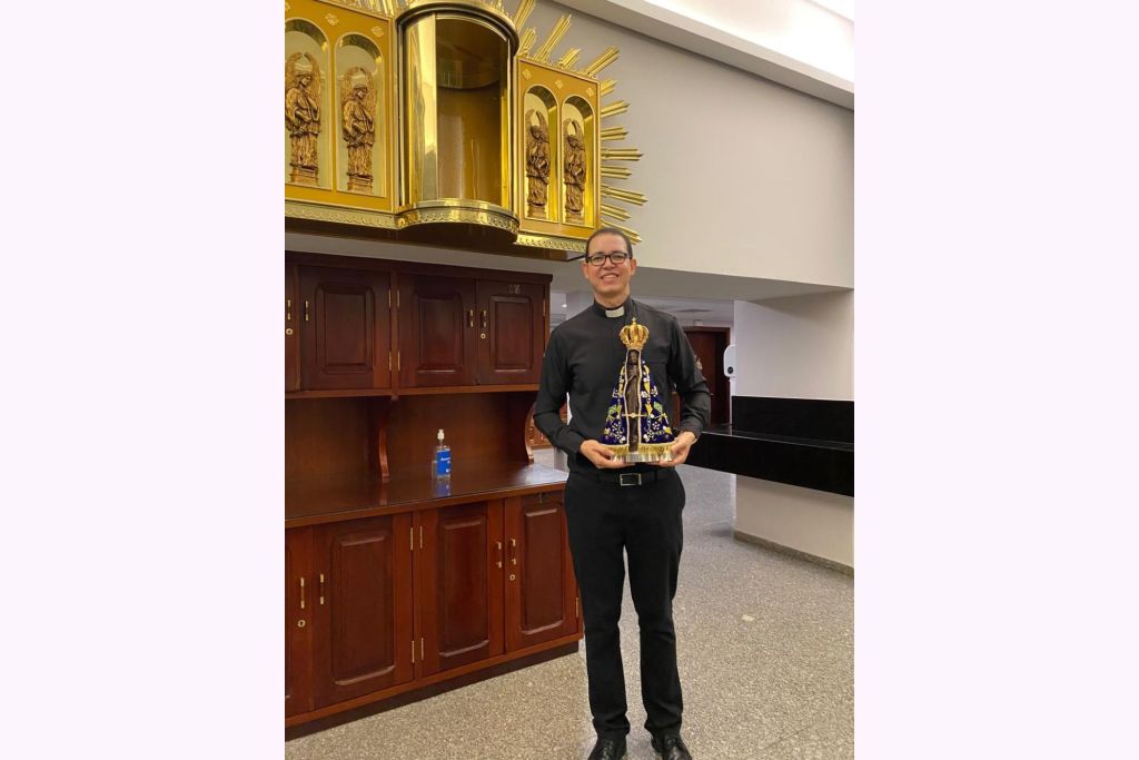 Fotografia de um jovem padre segurando uma imagem religiosa em um ambiente interno, com decoração dourada ao fundo, simbolizando fé e espiritualidade. Festa e Novenário em honra a Nossa Senhora Aparecida começa nesta sexta-feira em Paranaguá